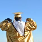 Graduate in gold gown celebrates achievement beneath clear blue skies.