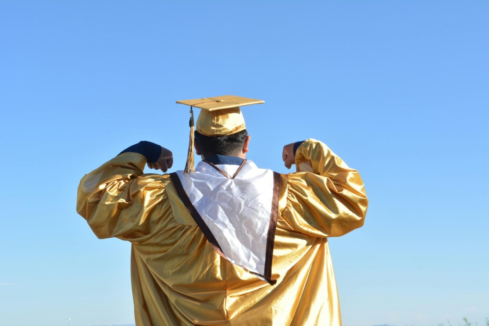 Graduate in gold gown celebrates achievement beneath clear blue skies.